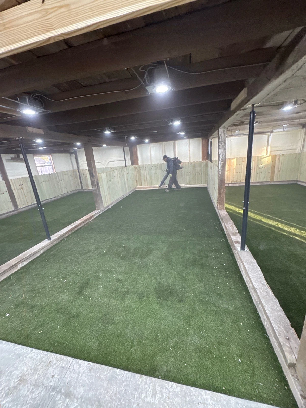 Indoor basement dog park with K9Grass Elite artificial turf, wood panel walls, recessed lighting, and blue steel posts in Pittsboro, IN.