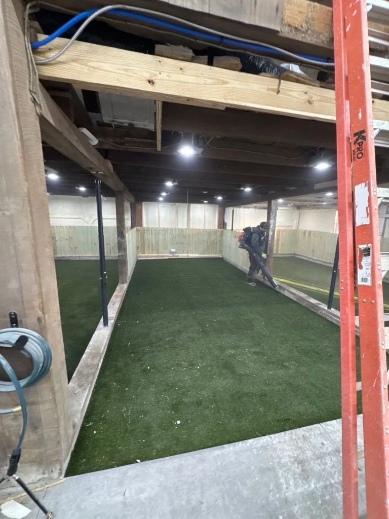 Indoor basement dog park with K9Grass Elite artificial turf, wood panel walls, recessed lighting, and worker using leaf blower during installation.