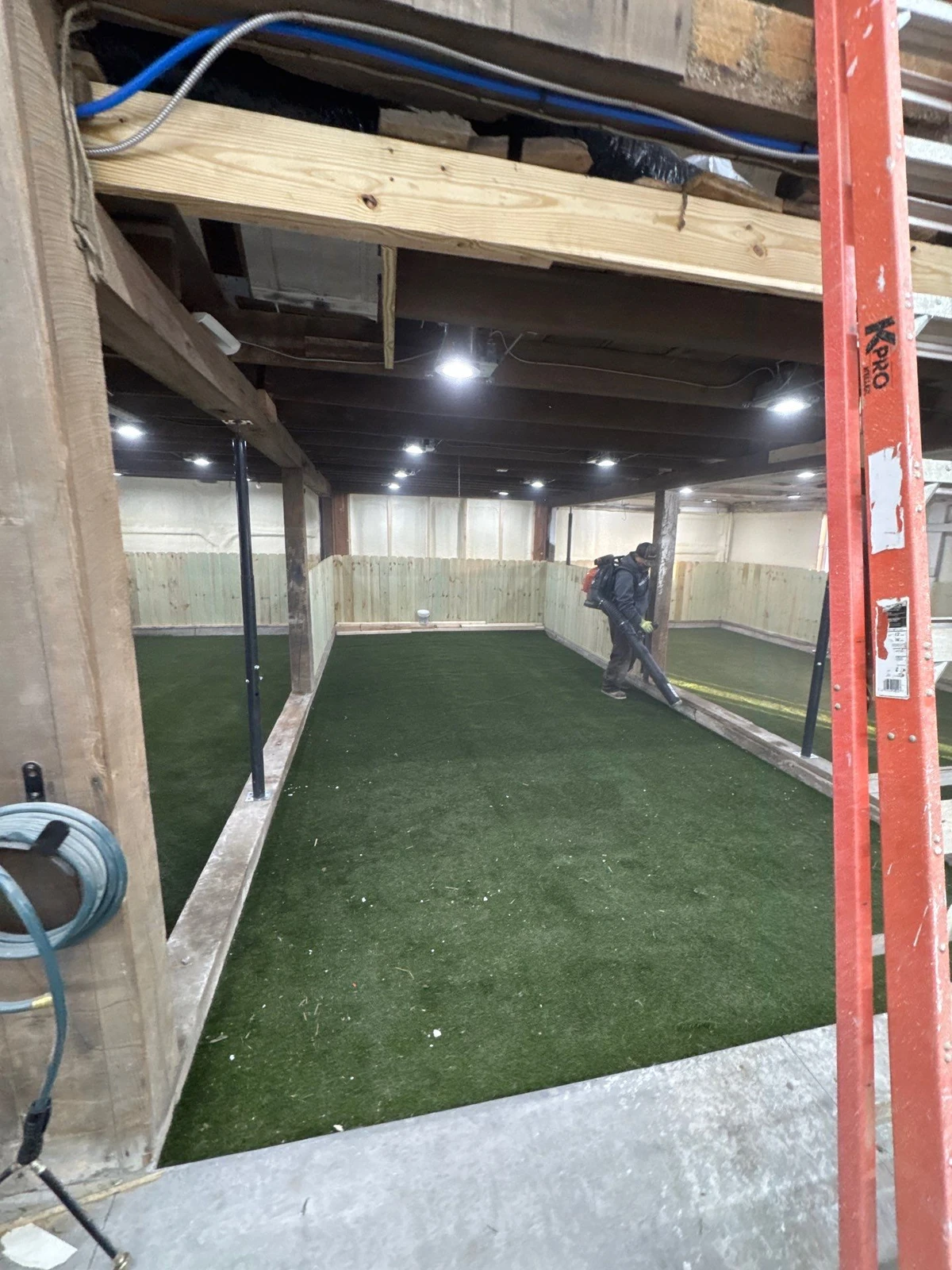 Indoor basement dog park with K9Grass Elite artificial turf, wood panel walls, recessed lighting, and worker using leaf blower during installation.