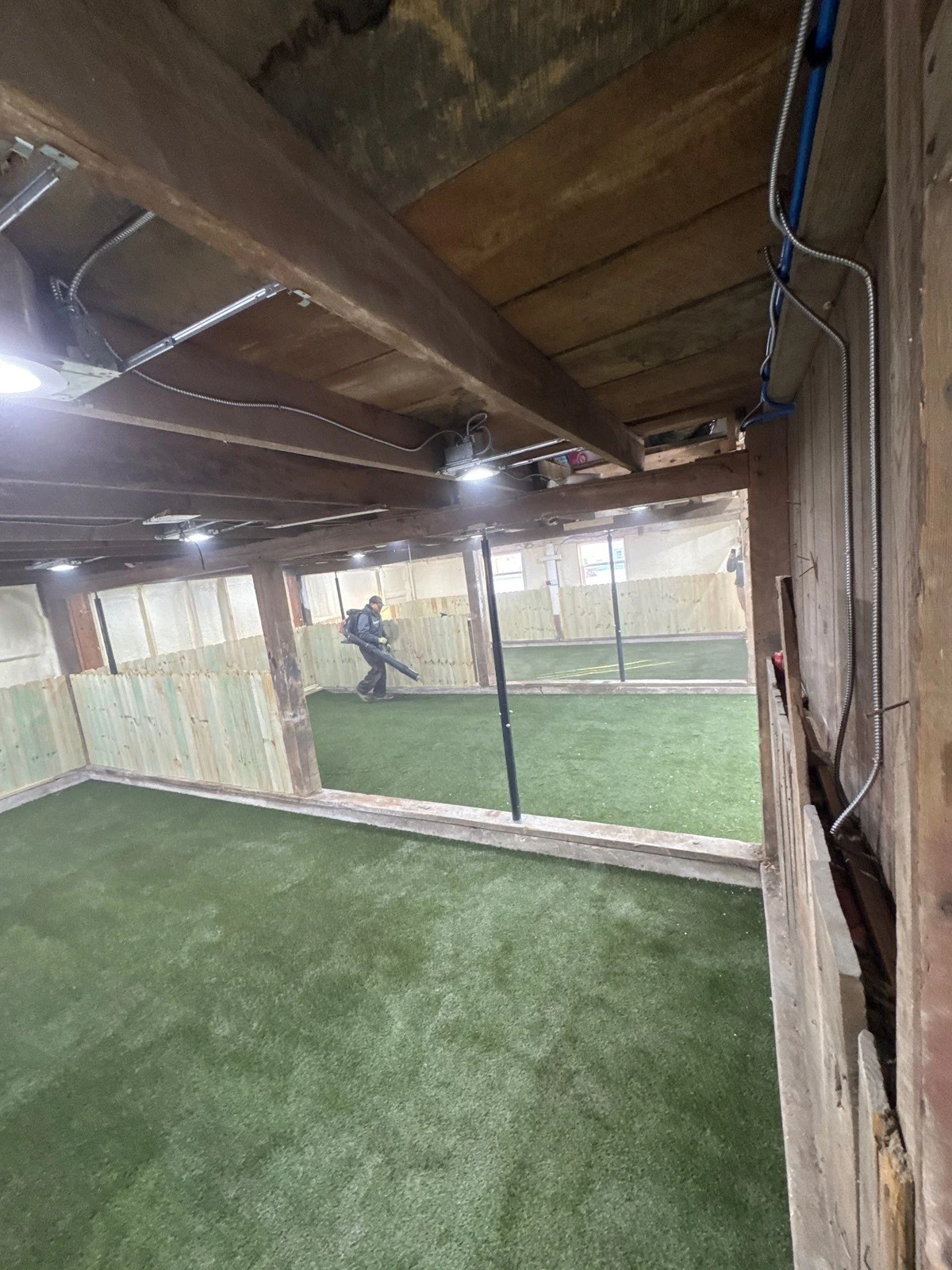 Indoor dog park under construction with K9Grass Elite artificial turf, exposed wood ceiling, conduit wiring, and worker using leaf blower.