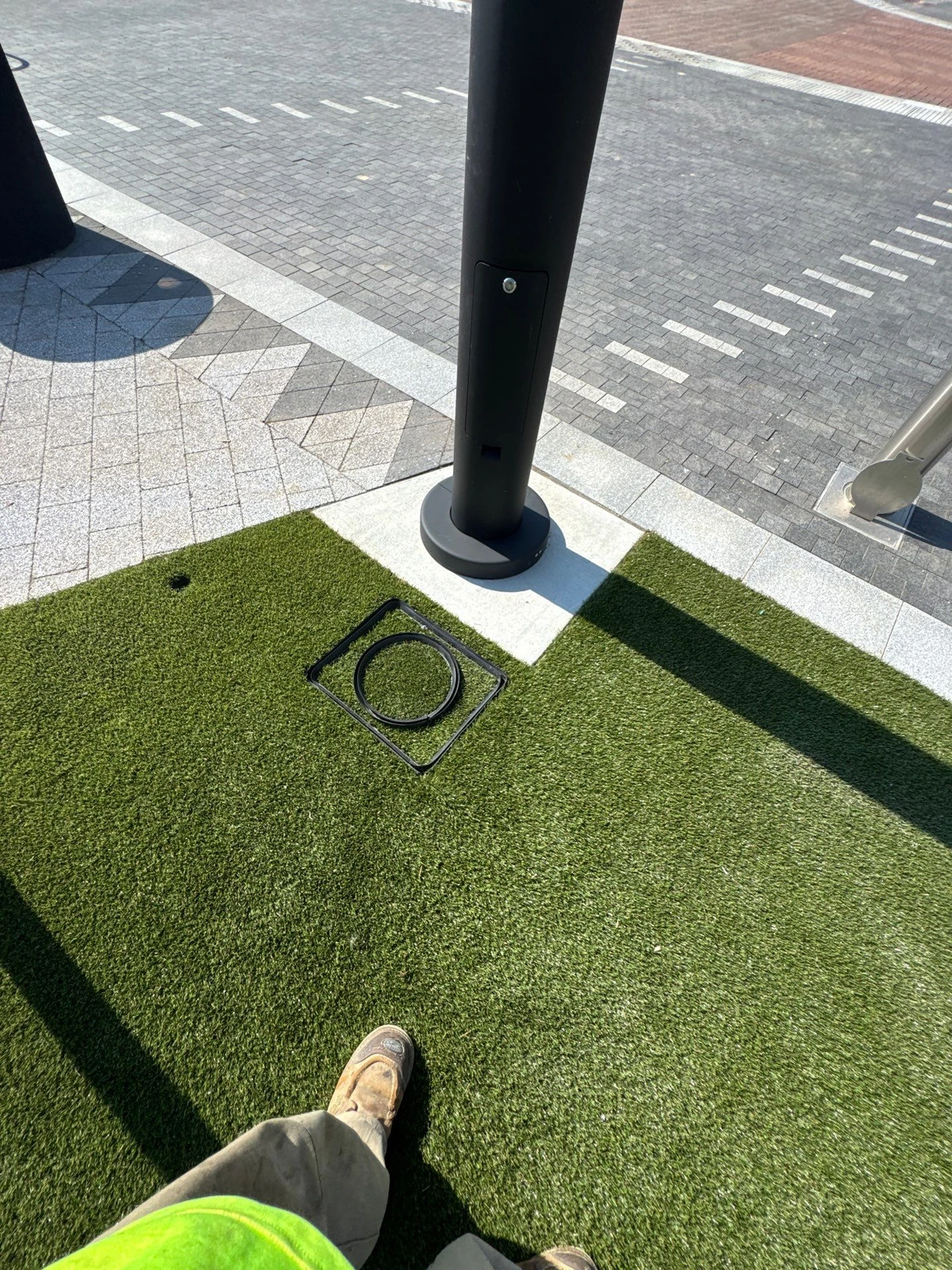 K9Grass artificial pet turf installed around black pole base with access port cutout, adjacent to decorative brick pavers.